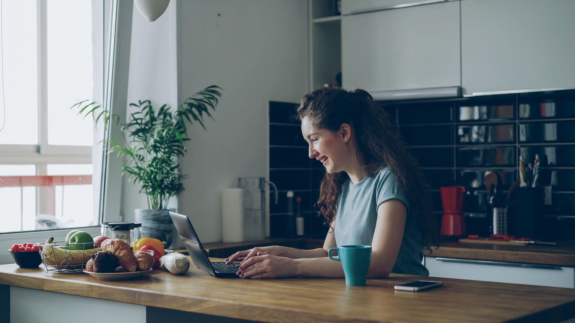 A woman at her kitchen table working through a job advert on her laptop, phone beside her
